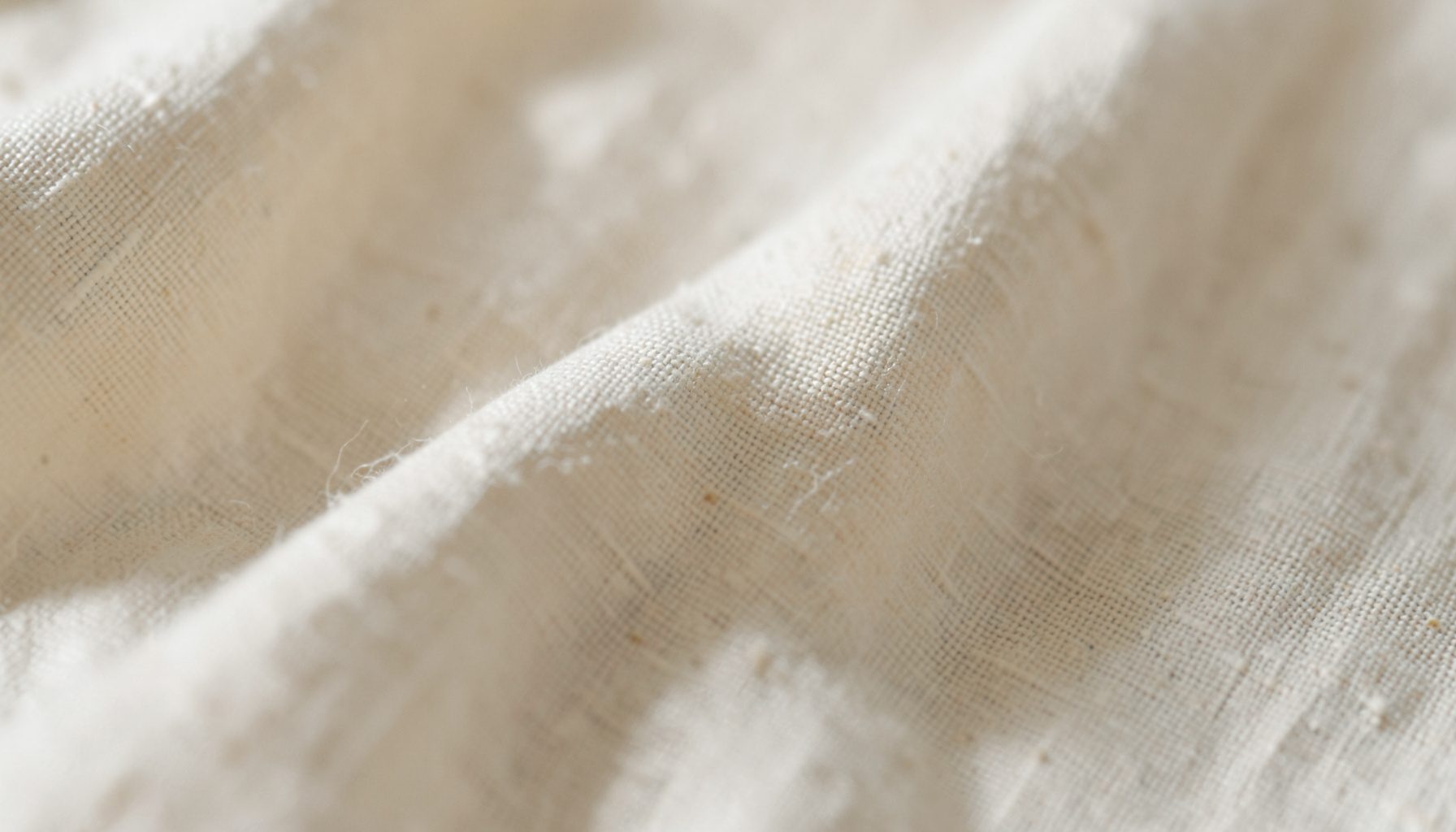Extreme close-up of pale linen fabric with a soft warm cream tone, gently folded to reveal subtle shadows and a quiet contemplative mood in muted ivory and sand hues