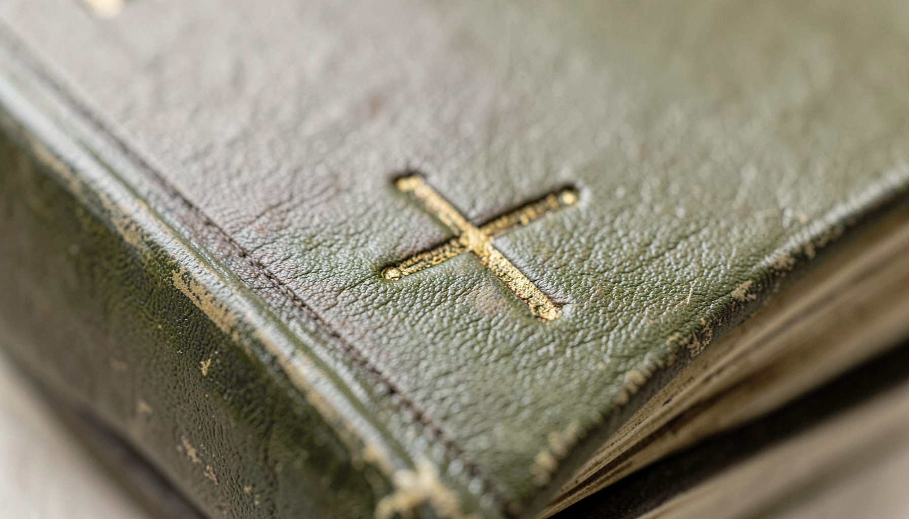 An extreme close-up of weathered olive-green leather binding with a faint gold-tooled cross, in deep taupe and cream tones, evoking a quiet, scholarly atmosphere.